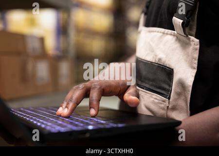 Close up of depot employee in workwear checking cargo parcels on racks using a laptop, fulfillment center operations for efficient merchandise distribution for e-commerce large business. Stock Photo