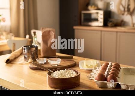 Kitchen countertop displaying carton of eggs, bowl of flour with spoon, butter on plate, glass bottles, rolling pin, paper bag, metal sifter, two glass bowls, blurred oven in background Stock Photo