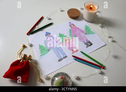 Flat lay winter family drawing on paper with colored pencils, candle, decorative lights, red gift pouch, and snow globe. Photo at an angle. Cozy holid Stock Photo