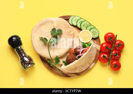 Wooden board of delicious pitas with falafel and fresh vegetables on yellow background Stock Photo