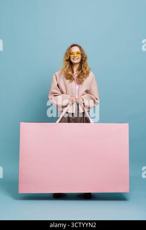 Happy shopper holding oversized pink bag against blue studio backdrop, ideal for Black Friday, discounts, retail ads, banners, and social campaigns. Stock Photo