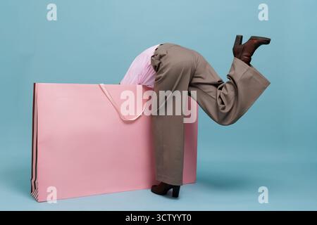 Playful woman dives into an oversized pink shopping bag on a blue backdrop with copy space, perfect for Black Friday and retail promotions. Stock Photo