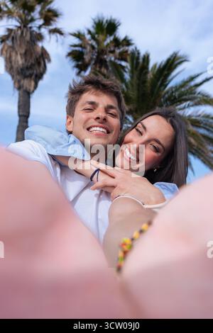 Playful young couple taking selfie with yellow phone in seaside palm lined park Stock Photo