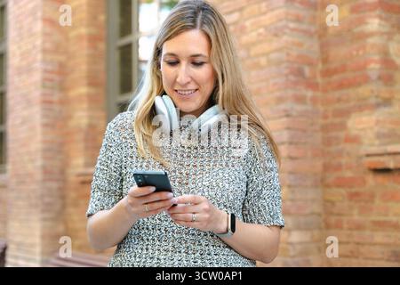 Young woman with headphones smiling while using smartphone outdoors happily Stock Photo