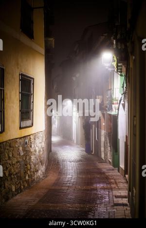 A deeply atmospheric photograph capturing a narrow, cobbled alleyway in Villajoyosa on a foggy night. The warm, yellow light from a street lamp illumi Stock Photo