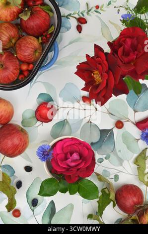 Red apples and roses on a tablecloth painted with green leaves. Harvest ready to eat or to process into preserves. Stock Photo