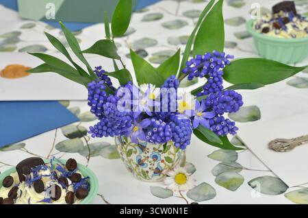 Little bouquet of grape hyacinths and daisies in regional vase on tablecloth painted with leaves. Envelopes and cream cupcakes. Stock Photo