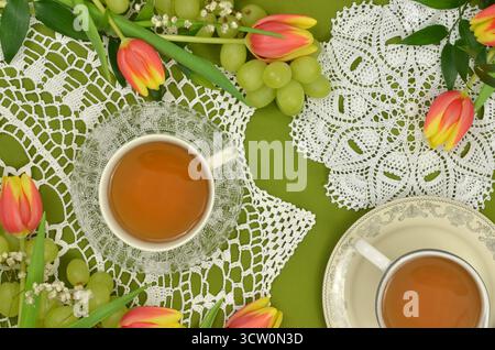 Five o'clock tea on handmade doilies, two cups of tea on green background with tulips, grapes and white gypsophila. Stock Photo