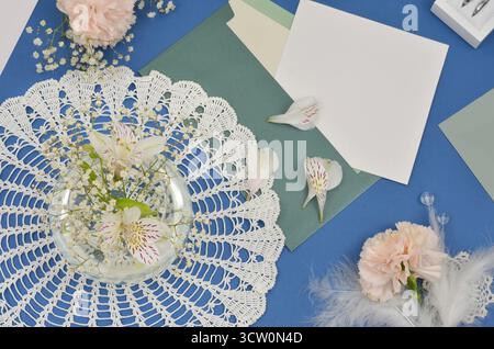 White, crochet doily decorated with light pink flowers and gypsophila flower, green envelope and white, empty paper to fill with content on dark blue. Stock Photo