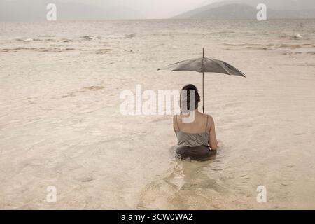 Woman from behind sitting in the sea water with a black umbrella under a cloudy and windy sky Stock Photo