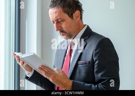 Mature businessman in formal suit with red necktie reading book by office window, concept of knowledge and learning. Selective focus. Stock Photo