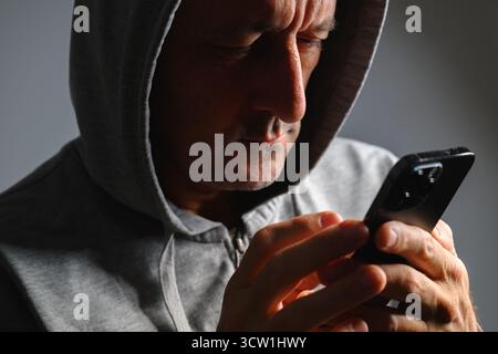 Mature man wearing hoodie using smartphone in low key light, concept of privacy, anonymity, or online communication. Selective focus. Stock Photo