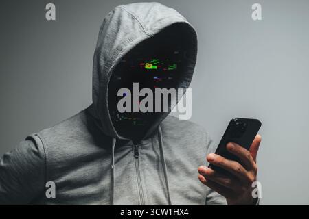 Mysterious hooded man stands in darkness with smartphone, face erased by glitch, representing anonymity and digital danger. Selective focus. Stock Photo