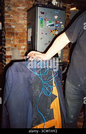 Analog flash photo of a dirty man in black holding a glowing blue LED strip. A grey fridge and a blazer on a chair frame the raw, surreal interior sce Stock Photo
