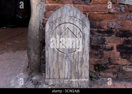 Weathered stone memorial with arched top and ancient script, set against a mossy red brick wall. Cracked gray stone and uneven ground convey historic. Stock Photo