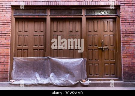 Brick building facade with three large wooden double doors, intricate panels, and weathered textures, partially covered construction materials Stock Photo