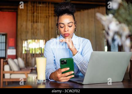 African american woman with a phone and laptop in a cozy space feeling unengaged while waiting Stock Photo