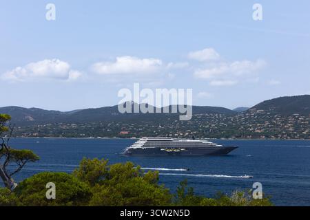 A black cruise ship is anchored in the blue sea off a hilly coastline. The sky is blue with white clouds. Stock Photo