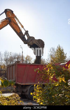 excavator pours concrete into a red truck under a clear blue sky on a construction site. Autumn trees surround the area, adding to the seasonal atmosp Stock Photo