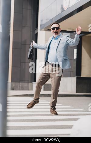 Businessman in a modern city confidently strides across a busy street crossing wearing a smart suit and stylish blazer while smiling Stock Photo