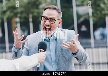 Confident businessman speaks passionately on a city street during an interview wearing a modern suit and glasses outside a bustling office in a Stock Photo