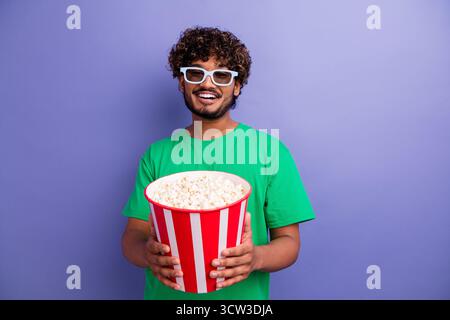 Positive man in casual green shirt holding striped popcorn bucket against violet background, enjoying leisure time and smiling Stock Photo