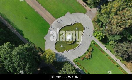 Aerial view of decorative fountain in city park. Drone shot of a symmetrical water fountain surrounded by green lawns, walking paths, and landscaped Stock Photo