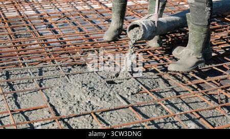 Concrete being poured over steel reinforcement mesh. Construction workers in boots pouring wet concrete onto a rebar grid, preparing the foundation Stock Photo