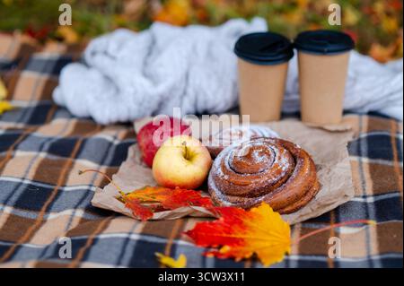 Autumn cozy picnic with a knitted sweater, plaid blanket, two paper cups of hot drinks, pastries, fresh apples, and colorful maple leaves falling arou Stock Photo