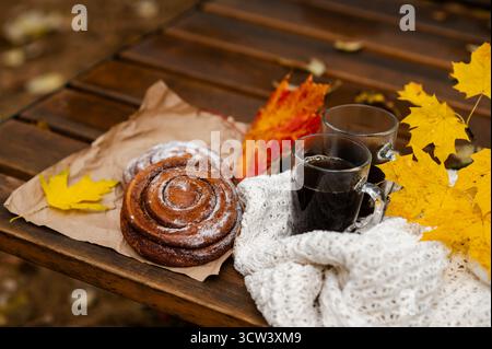 Two glass cups of hot coffee and cinnamon rolls with powdered sugar on craft paper, placed on a wooden bench decorated with yellow maple leaves and a Stock Photo