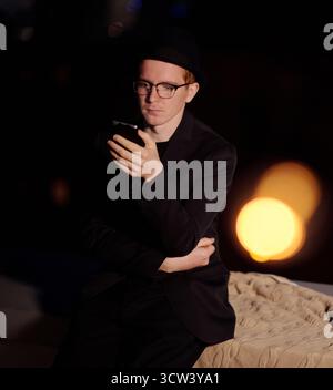 Young stylish Caucasian man in black outfit and glasses standing in the dark and checking social networks on smartphone, lens flare Stock Photo
