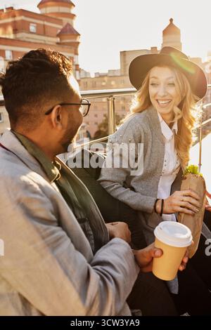 Young blond Caucasian pretty woman talking to her male friend and smiling cheerfully while sitting on rooftop terrace with drinks and food Stock Photo