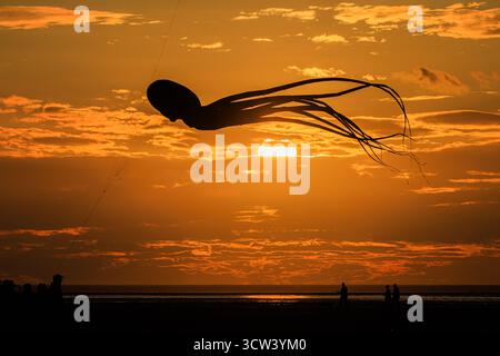 a striking silhouette of a large inflatable octopus kite with long trailing tentacles in vivid orange sunset cloudy sky reflected in calm sea Stock Photo