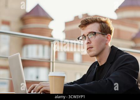 Young ginger-haired Caucasian man in glasses looking up thoughtfully while sitting at table in rooftop cafe and working on laptop Stock Photo