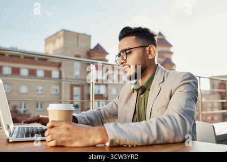 Young handsome Middle Eastern man with glasses sitting at table on rooftop terrace and working on laptop Stock Photo