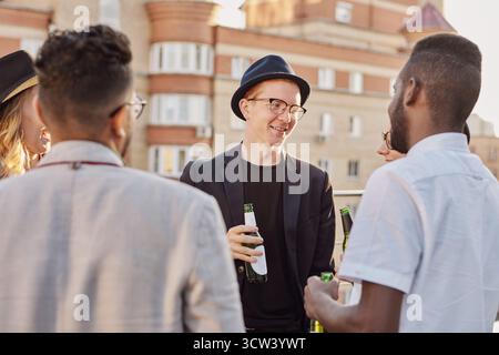 Multiethnic group of young stylish men and women standing on rooftop terrace, drinking beer and having friendly conversation Stock Photo