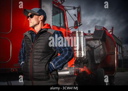 A truck driver stands with arms crossed, wearing sunglasses and casual clothing, next to a bright red truck in an urban area during a cloudy day. Stock Photo