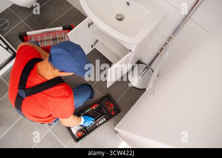 A repair technician is working on a bathroom sink, checking tools and plumbing under the cabinet in a modern home setting. Stock Photo