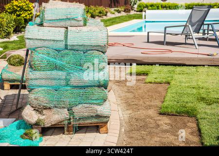 Freshly rolled sod is stacked and ready for installation next to a pool area in a backyard, showcasing preparation for landscaping on a sunny day. Stock Photo