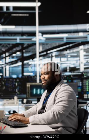 Upbeat data center IT expert uses PC to update servers tech, configuring gear using software. Jolly server room programmer reviewing hardware analytics, ensuring reliable network operations Stock Photo