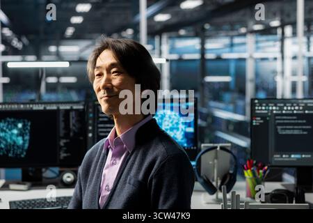 Smiling senior programmer in data center working on databases supporting AI applications. Happy Asian IT expert in server hub conducting experiments on artificial intelligence algorithms improvements Stock Photo