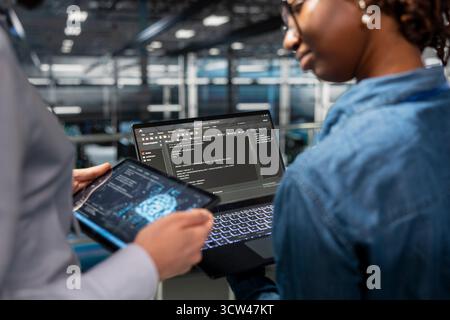 Close up of black technicians team reviewing LLM visualization with AI, overseeing machine learning outputs for the server farm operations. Engineers using tablet and laptop with intelligent systems. Stock Photo