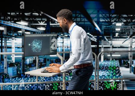 Black male specialist performing system maintenance on industrial platform in modern data center. IT expert using pc to do upgrades, resolving bugs and enhancing infrastructure stability. Stock Photo