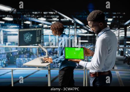 Black male technician using mockup screen on tablet to analyze neural network systems. System administrator uses green screen on tablet, reviewing LLM visualization on industrial platform. Stock Photo