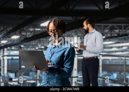 Black female programmer working on laptop in data center, handling maintenance tasks to ensure optimal performance for server automation. Woman IT expert checking code on laptop. Stock Photo