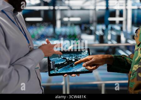 Military technician reviewing tactical dashboard on tablet screen in server hub, standing on industrial platform. Army expert using strategic communications and infrastructure software systems. Stock Photo