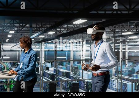 Black engineer using VR headset in data center to update server tech, configuring equipment using software tools. Server hub employee reviewing hardware analytics using virtual reality. Stock Photo