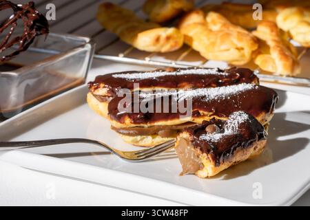 Three chocolate-glazed eclairs arranged on a white plate, captured in warm lighting for a cozy, appetizing presentation. Stock Photo
