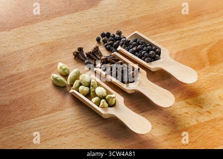Three common spices cardamom,clows,and black peppercorns. each asmall wooden scoop Isolated on Wooden Background Stock Photo