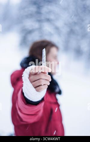 Woman is holding icicle towards the camera. Female model out of focus, while wearing winter red jacket standing in winter snow land. Stock Photo